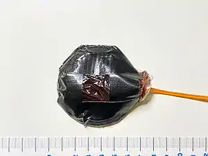 An assembled bullet hit squib device filled with approx. 25&nbsp;g of fake blood contained in a plastic pouch, secured onto a base with duct tape.