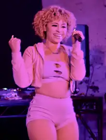 A young woman with frizzy dyed-orange hair holds a mic and smiles at a performance.