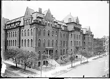 An elevated view of a five-story building on a street corner that takes up a long portion of a block. The building has multiple entrances leading to the sidewalk on the right. Trees line the street rising to the height of about two stories.