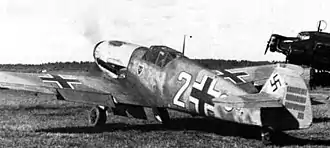 A black and white photograph a propeller driven fighter aircraft viewed from the rear-left. The aircraft is on a grass field, engine appears to be running. It bears three black and white crosses, two on the upper wings and one on the left side of the fuselage besides a large number "2". The tail rudder shows a black swastika plus rudder bears approximately 33&nbsp;small vertical black lines arranged in three groups of varying length.