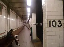 View of a narrow section of the southbound platform. There is a bench and wall to the left, as well as pillars with the number "103" to the right. There are subway tracks to the far right.